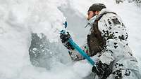 Ein Soldat im weißen Schneetarnanzug schaufelt Schnee mit einer blauen Schneeschaufel.