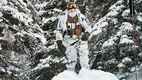 Ein Soldat im Schneetarnanzug steht mit seinen Skiern auf dem Iglu, dahinter verschneite Nadelbäume..