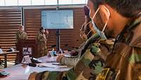 Mehrere Soldaten sowie Zivilisten sitzen an Tischen und schauen auf einen Bildschirm