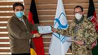 Ein Soldat und ein Zivilist halten gemeinsam ein Zertifikat in die Kamera, dahinter stehen drei Flaggen