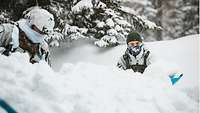 Zwei Soldaten in Schneetarnuniform schaufeln mit blauen Schneeschaufeln im weißen Schnee.