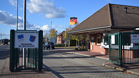 Wache Rechts Blick auf ein Wachhaus mit großem Dach, davor zwei Fahrbahnen für die Ein- und Ausfahrt