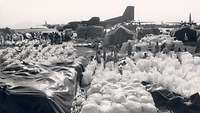 Lots of white sacks of relief supplies, behind them people, tents and airplanes