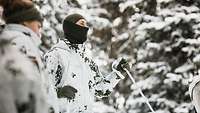 Zwei Soldaten in Schneetarnuniform stehen mit Skiern am verschneiten Waldhang.