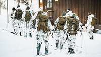 Soldaten in Schneetarnuniform laufen auf Skiern über den Schnee an einer braunen Hütte vorbei.
