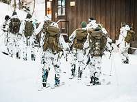 Soldaten in Schneetarnuniform laufen auf Skiern über den Schnee an einer braunen Hütte vorbei.
