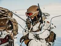 Ein Soldat in Schneetarnuniform mit Gefechtshelm hält den Hörer eines Funkgerätes an sein Ohr.