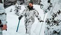 Drei Soldaten in Schneetarnuniform stehen am Waldhang, einer hält eine blaue Schneeschaufel.