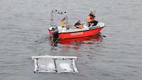 Kleines Motorboot mit zwei Feuerwehrleuten zieht zwei rechteckige, miteinander verbunde Matten durchs Wasser