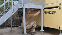 Soldat kniet unter einer Metalltreppe und überprüft die Podestanlage an dem angrenzenden Container
