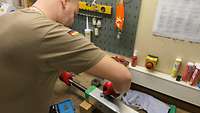 Ein Soldat steht an einer Werkbank. Vor ihm liegt eine Containerstütze aus Metall welche gewartet werden muss