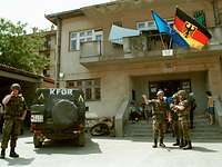 Deutsche Soldaten und ein KFOR-Militärfahrzeug vor einem Gebäude, an dem die Flagge der NATO und die Bundesdienstflagge wehen