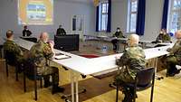 Acht uniformierte Soldaten sitzen in großen Abständen zueinander an Tischen in einem Konferenzsaal.