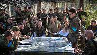 Mehrere Soldaten unterschiedlicher Nationen sitzen und stehen im Freien um eine Landkarte herum und besprechen sich