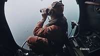 Ein Soldat sitzt an einer offenen Flugzeugtür und schaut mit einem Fernglas auf das Meer