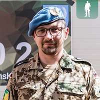 Ein Soldat mit blauem Barett steht vor einem Plakat mit grünem Polygon und lächelt in die Kamera