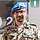 Ein Soldat mit blauem Barett steht vor einem Plakat mit grünem Polygon und lächelt in die Kamera