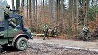 Bewaffnete Soldaten geduckt an einer Straße im Wald. Gesichert werden sie zusätzlich durch ein Fahrzeug mit Maschinengewehr.