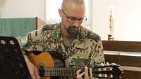 Ein Soldat spielt Gitarre in der Kirche des Camps, links im Bild ein Notenständer, rechts im Hintergrund eine große Kerze 