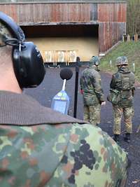 Ein Soldat mit Gehörschutz und einem Lärm-Messgerät steht auf einer Standortschießanlage hinter zwei Soldaten. 