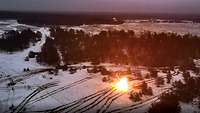 Auf einer verschneiten Landschaft steht ein schießender Panzer, im Hintergrund viele Bäume, mehrere Spuren verlaufen im Schnee