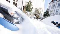 Ein Autoscheinwerfer lugt aus Schneemassen heraus. Im Hintergrund Soldaten beim Schneeschaufeln.