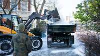 Ein Bagger kippt Schnee auf einen Laster. Davor steht ein Soldat.