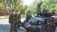 Mehrere Soldaten stehen um einen kleinen Panzer herum, zwei Soldaten stehen und hocken auf ihm.