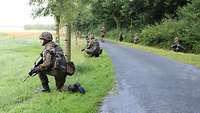 An einer Straße entlang hocken Soldaten und sichern mit ihren Waffen das Gelände ab.