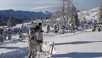 Soldaten im Schneetarnanzug stehen auf einer Schneefläche im Gebirge verteilt in einem Halbkreis um zwei Soldaten herum.
