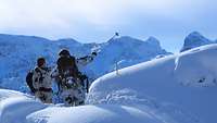 Zwei Soldaten im Schneetarnanzug stehen in tiefem Schnee, einer weist mit einem Skistock in eine Richtung.