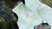 Ein Soldat hockt über einer Landkarte mit Markierungen. In der rechten Hand hält er einen Kompass.
