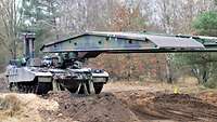 Ein Brückenlegepanzer der Bundeswehr fährt im Wald seine Brücke über Erdwällen aus.