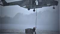 Per Seil nimmt ein fliegender Hubschrauber eine am Boden liegende Last auf, ein Soldat am Boden verbindet Seil und Last.