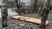 Am Ufer eines Flusses stehen vier Soldaten, in der Mitte liegt ein Floß aus Rundhölzern.