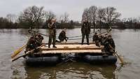 Eine Fähre, gebaut aus zwei Schlauchbooten und einer Holzplattform, schwimmt auf der Donau, darauf Soldaten mit Paddeln.