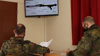 Zwei Soldaten sitzen an einem Tisch. Vor ihnen hängt oben ein Bildschirm, der das Gehäuse eines Maschinengewehrs zeigt.