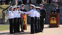 Sechs Marinesoldaten in Matrosenuniform halten ihre linke Hand über waagerecht gehaltene Fahne.