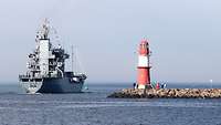 Ein graues Schiff in See, von hinten gesehen; rechts eine Hafenmole mit einem kleinen Leuchtturm.