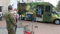 Ein Soldat und eine Frau mit Maske stehen vor einem Bus in Flecktarn. Darin ein weitere Soldat im medizinischen Kittel