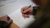 Ein Soldat hält in der rechten Hand einen Stift und schreibt etwas auf ein Blatt Papier 