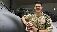 Ein Soldat mit einem American Football in den Händen lehnt an einem Hubschrauber und lächelt in die Kamera