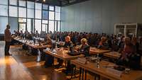 Ein Redner steht in einem großen Saal vor dicht besetzten Tisch- und Stuhlreihen 