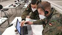 An einem Schreibtisch mit Laptop erklärt ein Soldat einem zweiten etwas am Bildschirm.