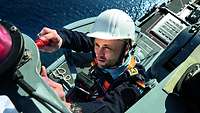 EIn Marinesoldat in blauer Arbeitsuniform und weißem Schutzhelm hoch in Aufbauten eines Schiffes.