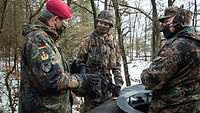 Ein Soldat mit roten Barett spricht mit zwei Soldaten, die neben und in einem Panzer stehen.