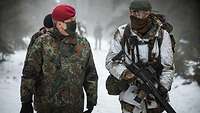 Ein Soldat in Flecktarn läuft neben einem Soldaten in Schneetarn durch den Wald.