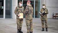 Ein Soldat hält eine Urkunde in der Hand. Zwei weitere stehen vor und neben ihm.