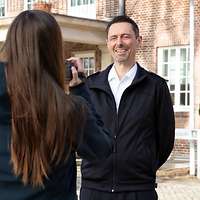  Kapitänleutnant Herms steht vor einem Gebäude und spricht in die Kamera. Die Kamerafrau steht mit dem Rücken zum Bild