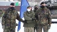 Drei Soldaten stehen nebeneinander mit gekreuztem Handschlag. Im Hintergrund steht ein weiterer Soldat mit der NATO-Flagge